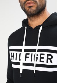 Uomo con barba che indossa un felpa nera con cappuccio, con stringhe bianche e un logo rettangolare bianco audace che mostra la scritta "HILFIGER" sul petto.