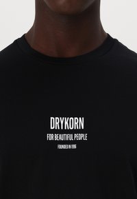 Must musta särk ümmarguse kaelusega, millel on valge trükitud tekst: "DRYKORN KAUNITE INIMESTE HEAKS" ja "ASUTATUD 1996".