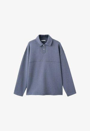 Junger Mann in lockeren beigen Hosen, braunen Schuhen und einem langärmligen blauen Polohemd steht vor einem schlichten weißen Hintergrund.