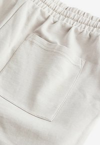 Tela de algodón beige claro con un acabado texturado, que cuenta con una cintura elástica y un bolsillo trasero único con detalles de costura visibles.