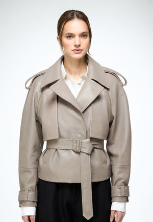 Kobieta ubrana w taupe skórzaną kurtkę z paskiem i szerokimi klapami na białej koszuli, stojąca na jednolitym białym tle.