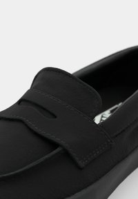 Sorte lærloafer med rund tå, med en dekorativ stropp og sømdetaljer. Glatt tekstur og minimalistisk design.
