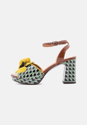Platform sandal with a tan ankle strap, green and white patterned base, yellow flower accent, and a chunky high heel.