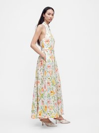 Femme debout dans une robe maxi fleurie sans manches avec poches latérales, portant des mules à talons bas de couleur claire, sur un fond uni.