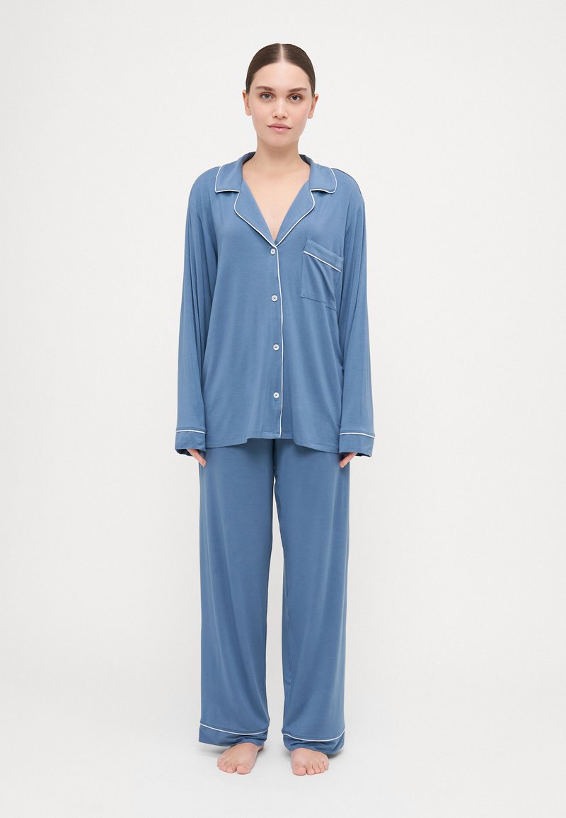 Blue pajama set in soft fabric featuring a collared shirt with buttons and a chest pocket, paired with loose-fitting pants and white piping details.
