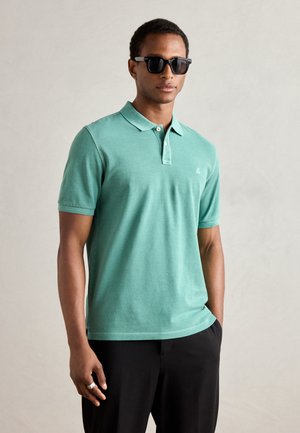 Camisa polo verde menta de manga corta, con cuello y tapeta de dos botones. Se lleva con pantalones negros y gafas de sol oscuras. Detalle de logo sutil.