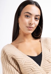 Cardigan bej din tricot cu un decolteu adânc în V, purtat peste un top negru, având o textură moale și mâneci late; cercei mari, rotunzi, vizibili.