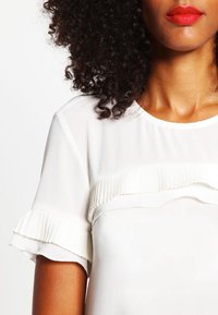 Bílá blůza vyrobená z měkké látky, s plišovanými detaily přes hruď a volánkovými krátkými rukávy. Hladká textura s volným střihem.