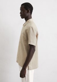 Camicia a maniche corte di colore beige a righe, realizzata in tessuto leggero, con colletto, taschino sul petto e chiusura con bottoni.