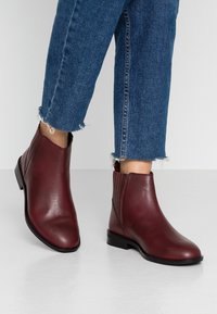 Mujer vistiendo botas de cuero rojo oscuro hasta el tobillo con paneles elásticos laterales y jeans azules con dobladillo deshilachado sobre un suelo y fondo blancos.