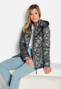 Grau gestepptes Jacket mit Kapuze, glänzendes Material und Reißverschluss. Dazu helle blaue Hose und ein weißes Hemd.