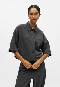 Graues, kurz geschnittenes Shirt aus strukturiertem Stoff mit spitzem Kragen, kurzen Ärmeln und einer Knopfleiste auf der Vorderseite. Verfügt über eine Taschenapplikation.