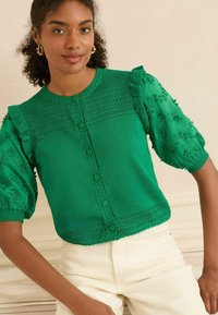 Blusa verde a maglia con design testurizzato, maniche a volant, ricamo floreale e chiusura frontale con bottoni. Abbinata a pantaloni di colore chiaro.