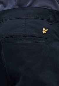 Pantalones de algodón azul marino con un bolsillo parche en la parte trasera que exhibe un logotipo de pájaro bordado en oro, mostrando una textura suave y un diseño ajustado.