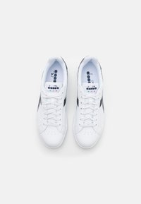 White athletic sneakers with black accents and perforations. Features flat laces and a logo on the tongue. Smooth synthetic material.