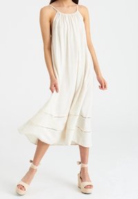 Beige Leinen-Maxikleid mit gerafftem Halterneck, ausgestattet mit subtilen horizontalen Nähten am Saum. Getragen mit beigen Espadrilles.