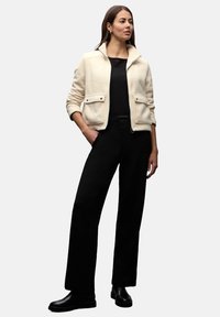 Kremowa marynarka z mieszanki wełny z kołnierzem stójką, przednimi kieszeniami, zestawiona z czarną bluzką i szerokimi spodniami, podkreślona czarnymi botkami.