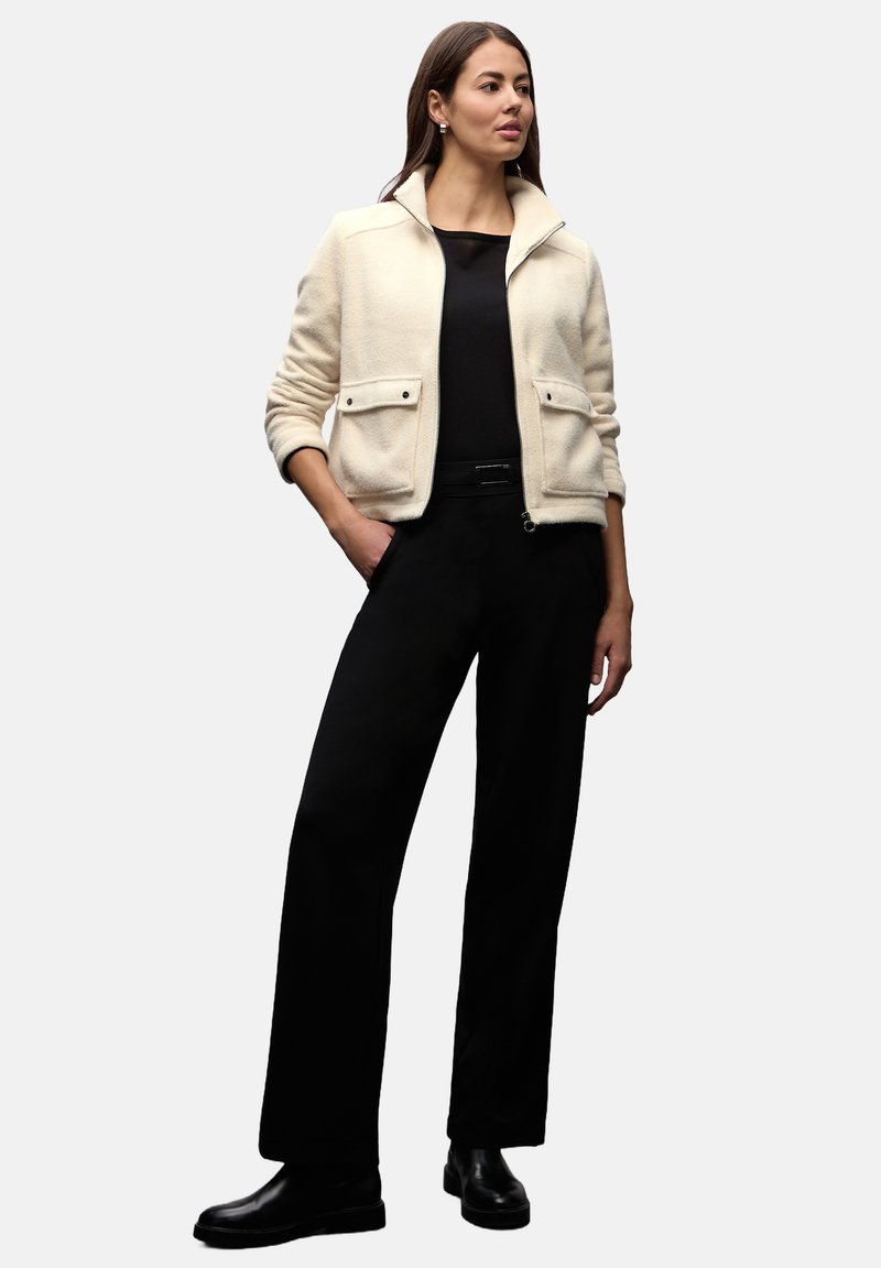 Kremowa marynarka z mieszanki wełny z kołnierzem stójką, przednimi kieszeniami, zestawiona z czarną bluzką i szerokimi spodniami, podkreślona czarnymi botkami.