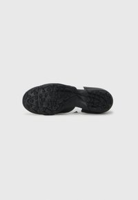 Suela de zapatilla deportiva negra con un diseño texturizado, que presenta una franja blanca suave en el centro y patrones de agarre visibles.