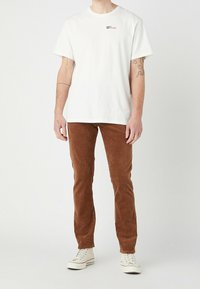 T-shirt en coton blanc à manches courtes, avec un petit logo, associé à un pantalon en velours côtelé marron et des baskets blanches.