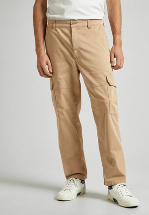 Pantalones cargo beige de tejido duradero con bolsillos laterales, diseño de pierna recta y cinturilla estándar. Llevados con zapatillas blancas.