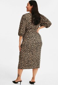 Leopardenmusterkleid aus beigem und schwarzem Stoff, mit Dreiviertelärmeln, taillierter Taille und mittellanger Länge bis zur Wade. Schwarze High-Heels mit offenem Zehen.