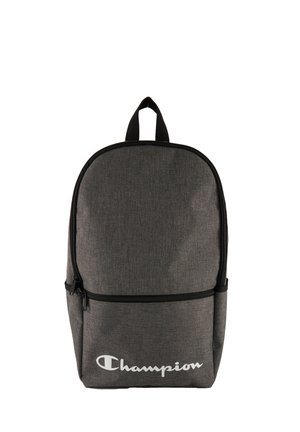 Mochila de tela gris con forma redondeada, que cuenta con un bolsillo delantero con cremallera y un asa de transporte en la parte superior. Logo blanco prominente de "Champion" en la parte inferior.