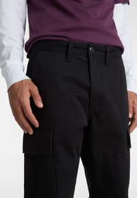 Schwarze Cargo-Hosen aus Baumwolle mit Seitentaschen, Knopfverschluss und geradem Schnitt. Getragen mit einem lila Hemd darüber.