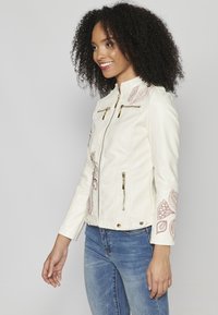 Veste en simili cuir blanc avec quincaillerie dorée, poches zippées et broderies florales sur les manches. Design ajusté et longueur cropped.