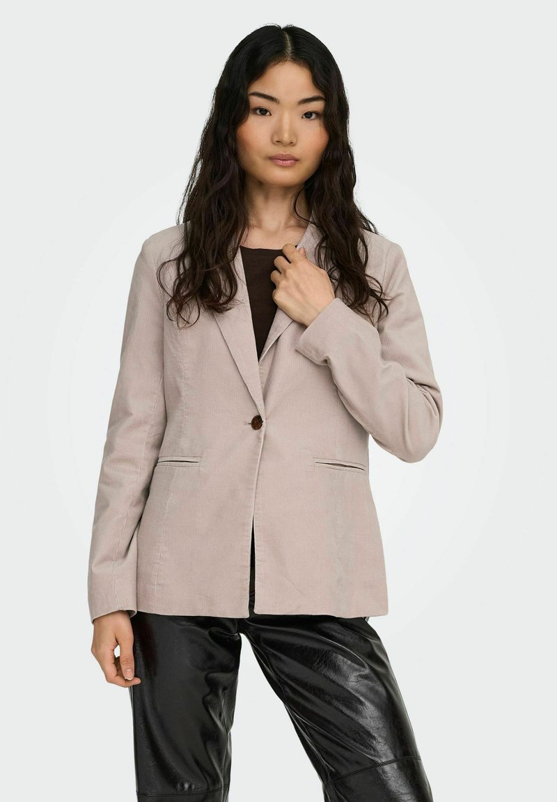 Jasnobeżowa marynarka z teksturowanego materiału z pojedynczym zapięciem na guzik, klapami z wcięciem oraz dwoma przednimi kieszeniami, zestawiona z czarnymi spodniami.