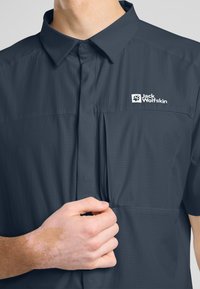 Korte-mouwen overhemd met knopen in donkerblauw, van een gestructureerde stof met een borstzak; heeft een wit logo. Hand die de stof vasthoudt.
