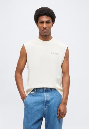 Hombre con camiseta blanca sin mangas con pequeño logo en el pecho izquierdo y vaqueros azules, de pie con las manos en los bolsillos contra un fondo blanco.