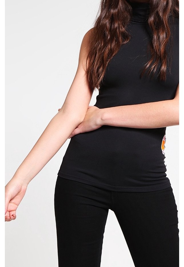 Femme portant un haut noir sans manches à col roulé et un pantalon noir, aux longs cheveux bruns, ajustant son bras gauche sur un fond blanc.