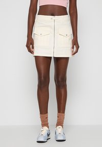 Minifalda de mezclilla blanca con cremallera frontal, dos grandes bolsillos de parche y detalles de costura visibles; combinada con zapatillas azul claro y calcetines marrones.