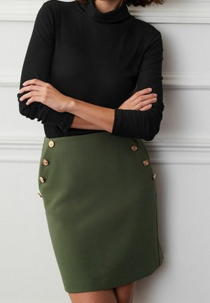 Femme les bras croisés, portant un haut noir à manches longues et une jupe vert olive ornée de boutons décoratifs dorés, debout contre un mur blanc.