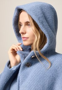 Abrigo azul con capucha, hecho de un tejido texturizado, que presenta un alto cuello, dos grandes botones y un aspecto suave y cálido.