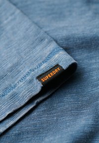 Lyseblåt stof med en struktureret finish, der har et sort mærke med orange "SUPERDRY" tekst på kanten. Blødt, afslappet design.