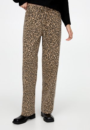 Persoon die hooggetailleerde, loszittende broek met luipaardprint draagt, zwarte laarzen en een zwarte top met lange mouwen, staand tegen een witte achtergrond.