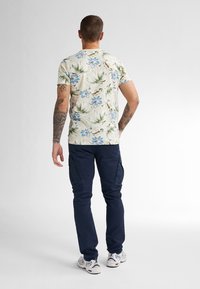 Katoenen t-shirt in lichtbeige met blauw bloemenpatroon en groene bladeren; gecombineerd met donkerblauwe cargobroek en witte sneakers.