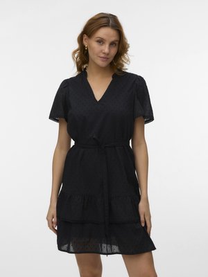 Γυναίκα με κυματιστά μαλλιά που φοράει μαύρο φόρεμα με κοντά μανίκια, V-λαιμόκοψη, ζώνη στη μέση και φούστα με βολάν, στέκεται μπροστά σε λευκό απλό φόντο.