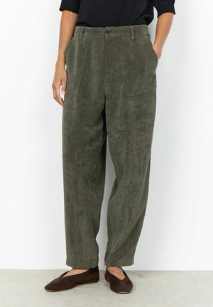 Person wearing loose, olive green corduroy pants with front pockets and brown flat shoes, standing against a plain white background.
