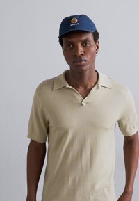 Beige kortærmet polo skjorte med flad krave og en subtil tekstur, parret med en blå cap med et gult logo.