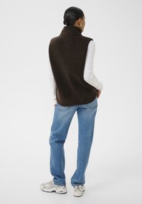 Mujer con el cabello oscuro recogido, vistiendo un chaleco de forro polar marrón sobre una camisa blanca de manga larga, jeans azules y zapatillas deportivas blancas, de pie mirando hacia atrás.