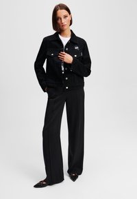 Črna denim jakna z dvema žepoma na prsih in gumbnim zapiranjem, stilizirana z belo srajco in ohlapnimi črnimi hlačami. Ima detajl z logotipom.