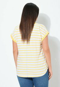 Lyhyet hihattava raidallinen toppi, jossa on keltaisia ja valkoisia värejä, pyöreä helma ja näkyvät saumat, yhdistettynä sinisiin farkkuihin.