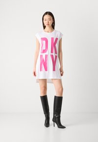 Abito bianco oversize con grande scritta rosa "DKNY". Abbinato a stivali neri al ginocchio con tacco a stiletto. Design semplice e moderno.