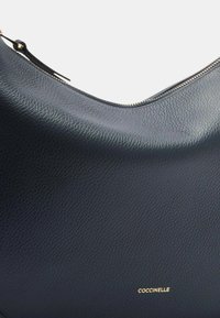 Navy leren handtas met een gestructureerde afwerking, gebogen vorm, gouden hardware en een ritssluiting. Merkennaam "Coccinelle" in het goud geperst.