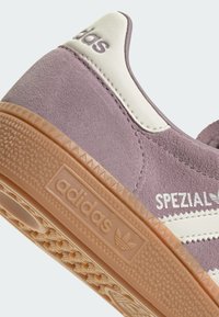 Ljubičaste suede tenisice s bijelim kožnim detaljem i gumenim potplatom. Sadrži Adidas brendiranje i teksturirani uzorak potplata.