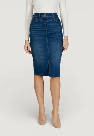 Donna che indossa una gonna a tubino aderente in denim blu scuro con una spacco frontale, abbinata a un top bianco a maniche lunghe e tacchi alti color nude.