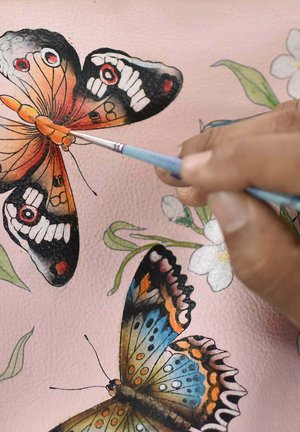 Schilderij van kleurrijke vlinders op een roze gestructureerd materiaal; fijne details omvatten levendige patronen en een hand die een kleine penseel vasthoudt.